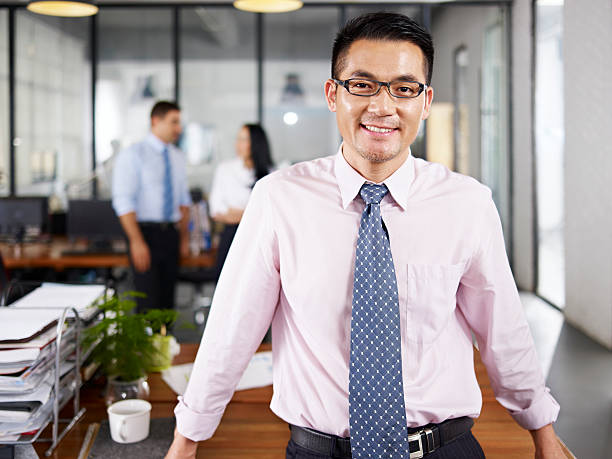 asian businessman standing in office happy and smiling with multinational colleagues talking in background.